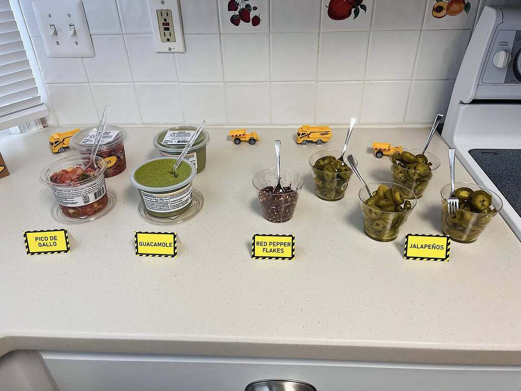 Table spread of taco bar condiments including pico de gallo, guacamole, red pepper flakes, and jalapeños.