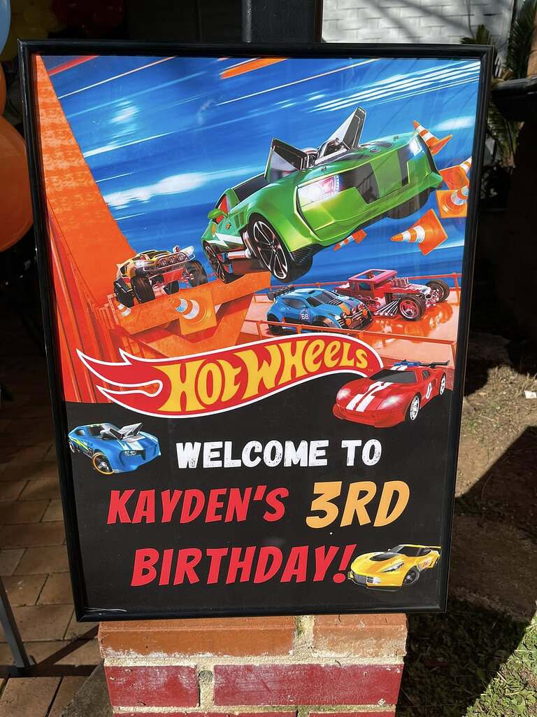 Hot Wheels themed party sign reading Welcome to Kayden's 3rd Birthday with race car graphics.