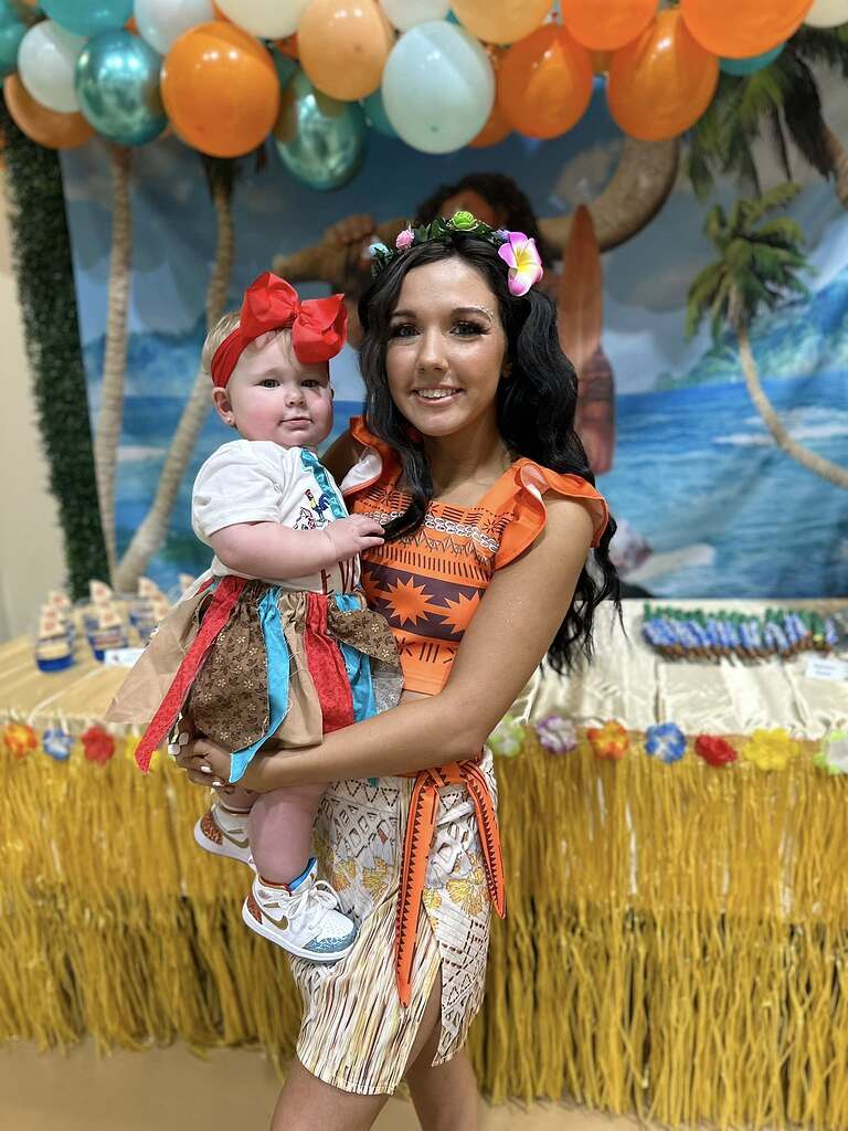 A woman dressed as Moana holding a baby wearing a matching tropical-themed outfit at a birthday party.