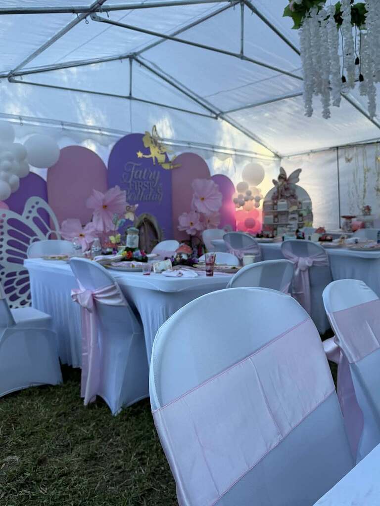 Fairy-themed first birthday party setup with purple and pink backdrop, floral decor, and linen-covered tables.