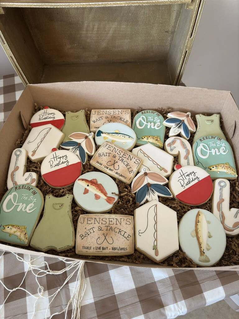 Box of fishing-themed sugar cookies for a first birthday party including fish, bobbers, and tackle boxes.