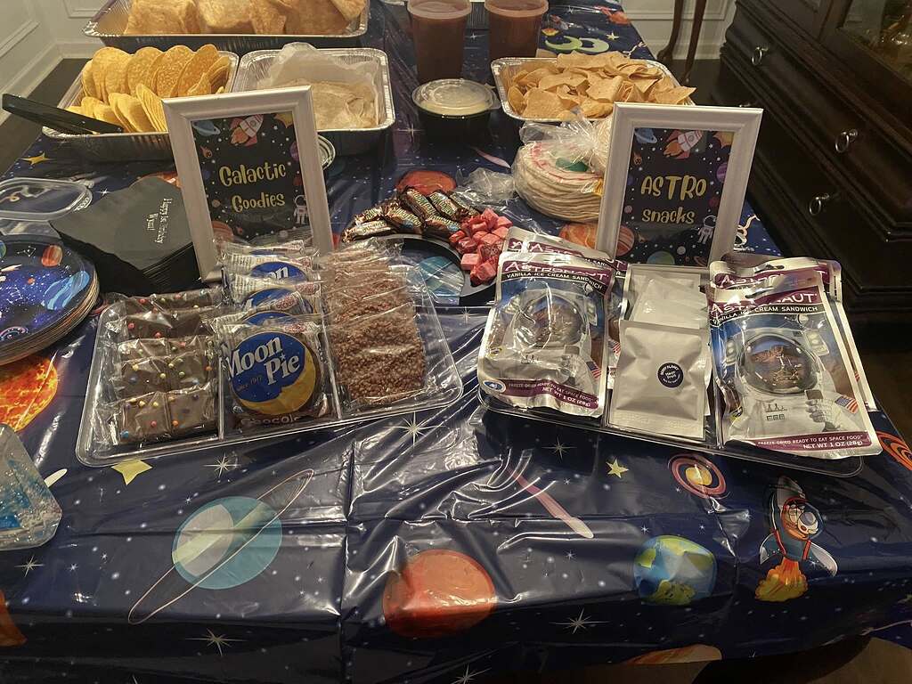 Space-themed party food table with Moon Pies, astronaut ice cream, and signs for Galactic Goodies and Astro Snacks.