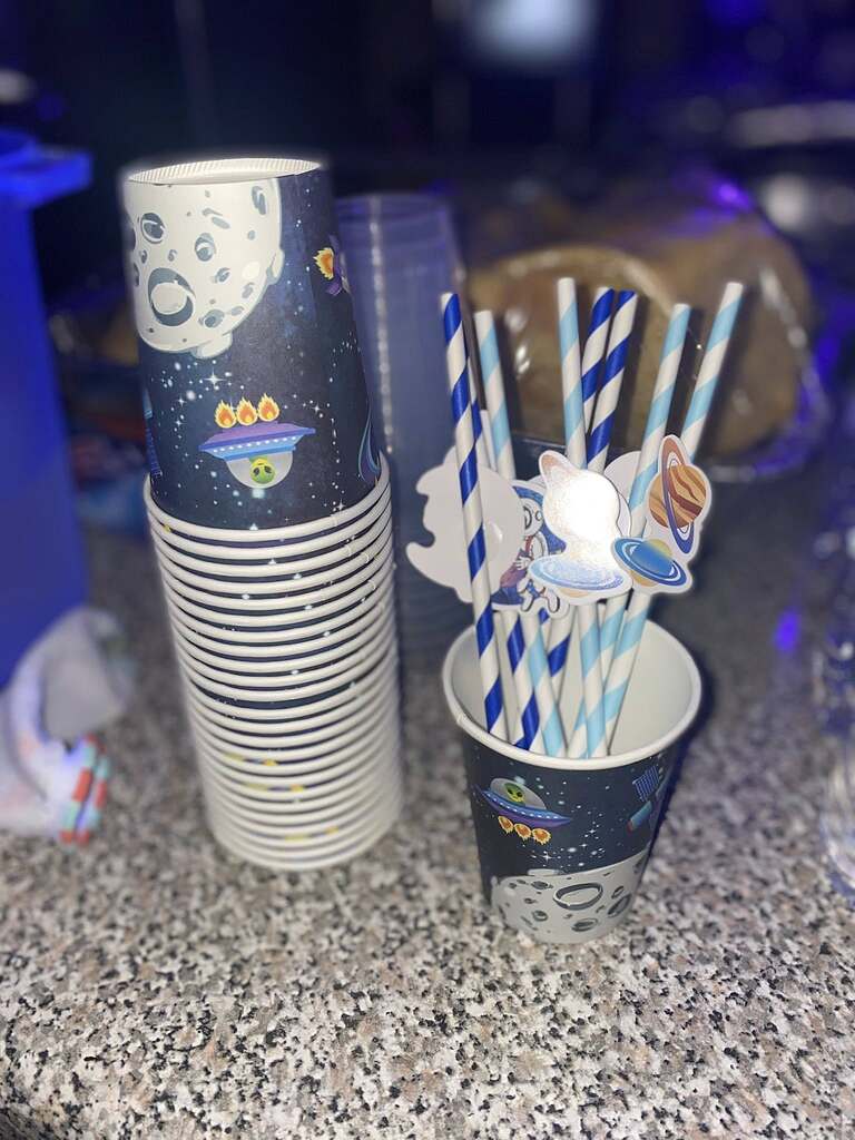 Stack of space-themed paper cups next to a cup filled with blue striped straws adorned with planet decorations.