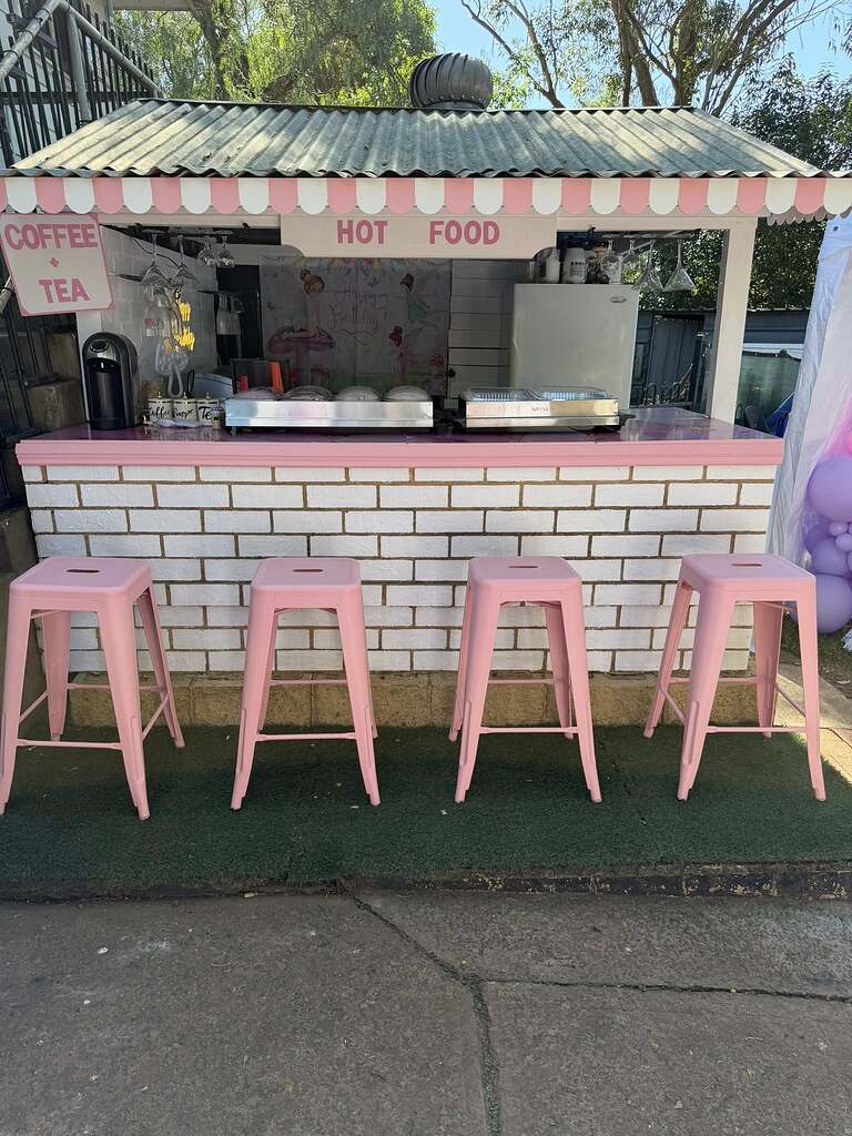 Outdoor catering food stall with a white brick base, pink trim, and four matching pink stools.