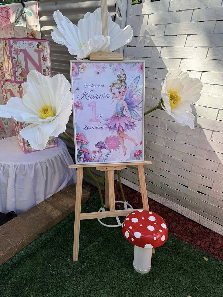 Easel displaying a fairy-themed sign for Kiara's 1st birthday party, surrounded by large paper flowers and a mushroom stool.