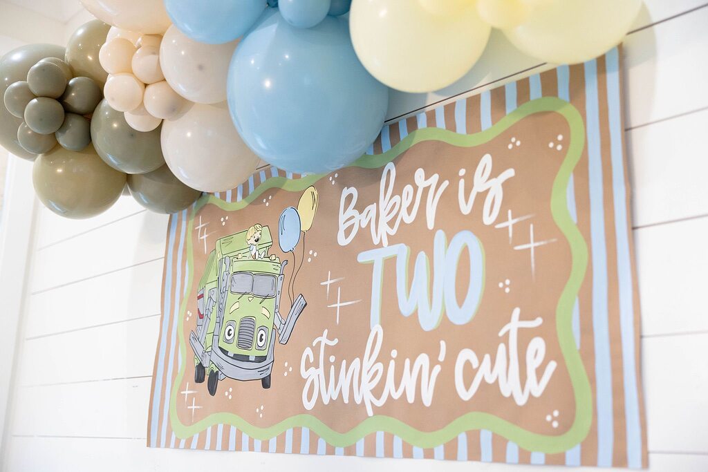 Baker is two birthday banner with a cartoon garbage truck illustration and a balloon arch background