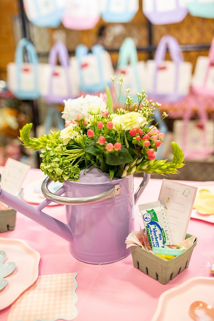 A lavender watering can filled with a festive floral centerpiece sitting on a pink table setting.