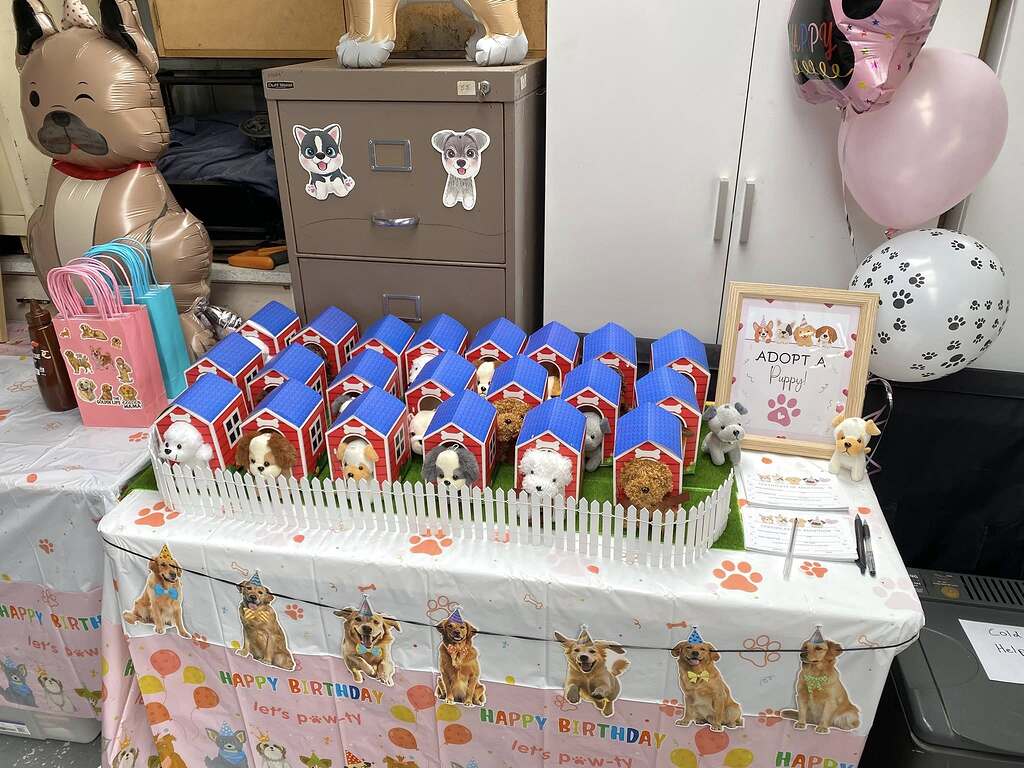 Puppy adoption birthday party display featuring plush dogs in small red kennel boxes behind a white picket fence