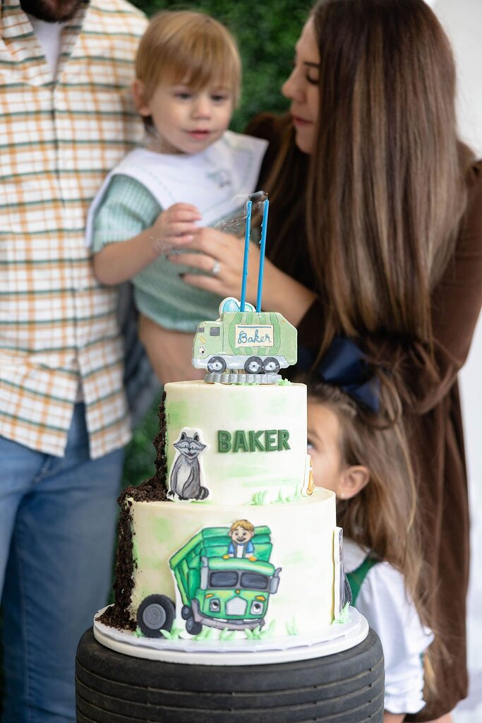 Two-tiered birthday cake with trash truck theme and Baker name label featuring a child being held by parents