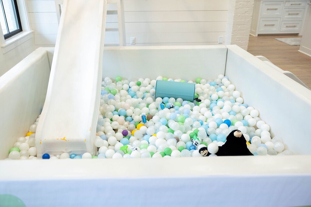Indoor white foam ball pit for kids with a small attached slide and toys.