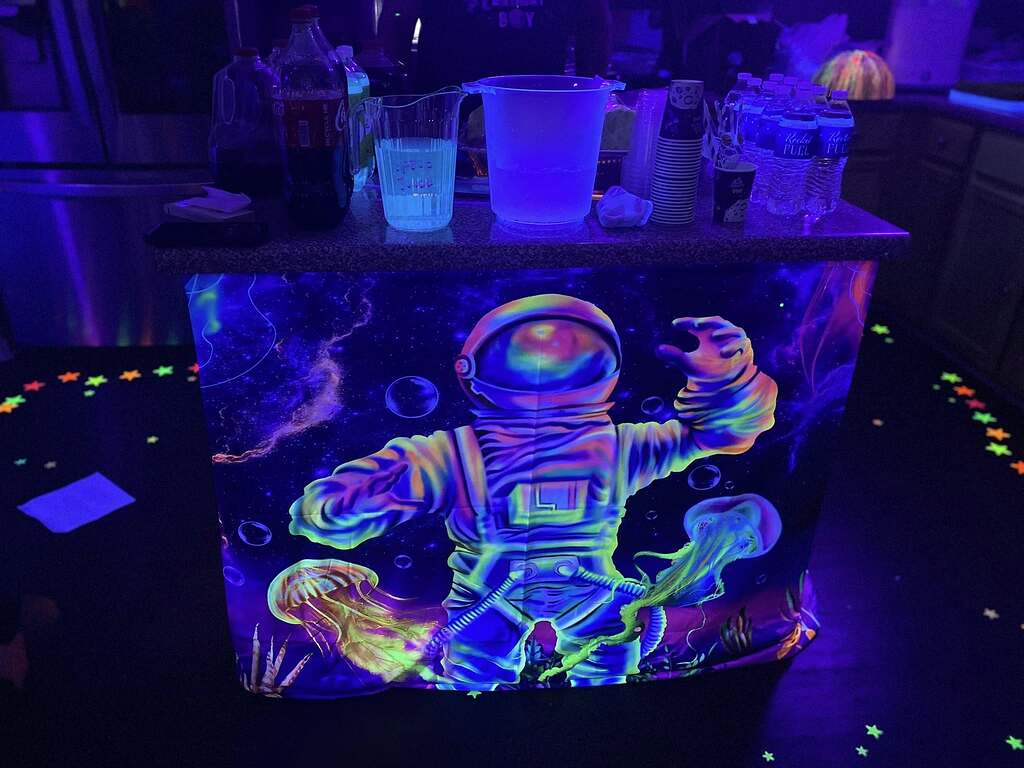 Table covered in a glowing neon astronaut and jellyfish tablecloth at a party with drink supplies on top.