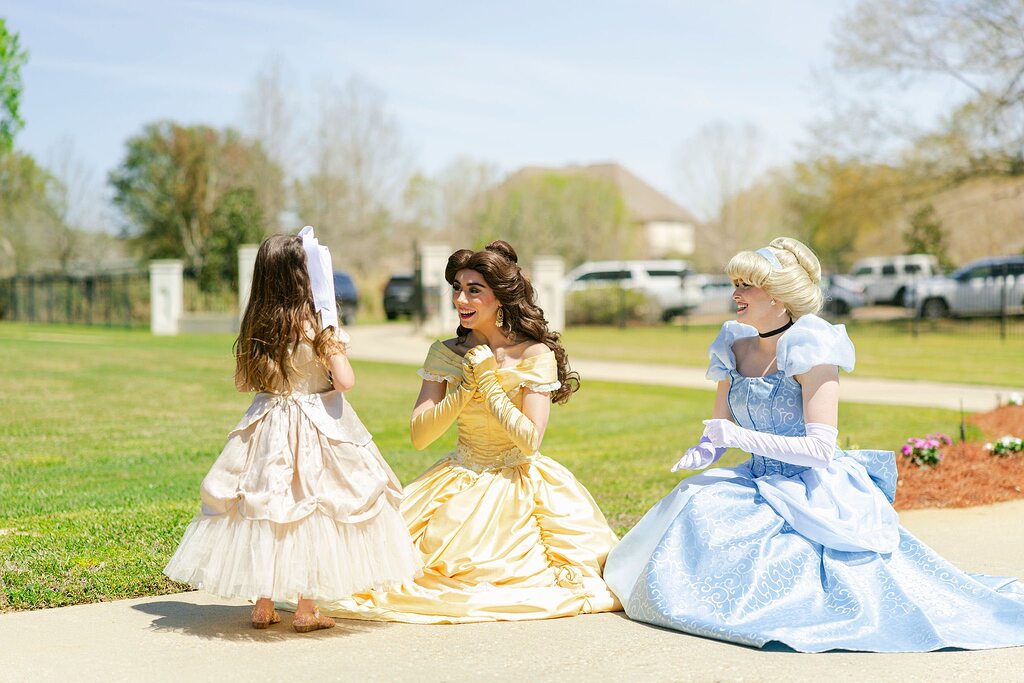 A young girl in a princess dress chatting with Belle and Cinderella character performers outdoors.