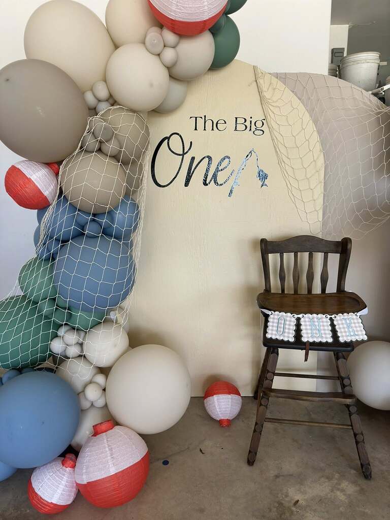 Fishing themed first birthday party backdrop with balloons, decorative nets, bobber lanterns, and a wooden high chair.