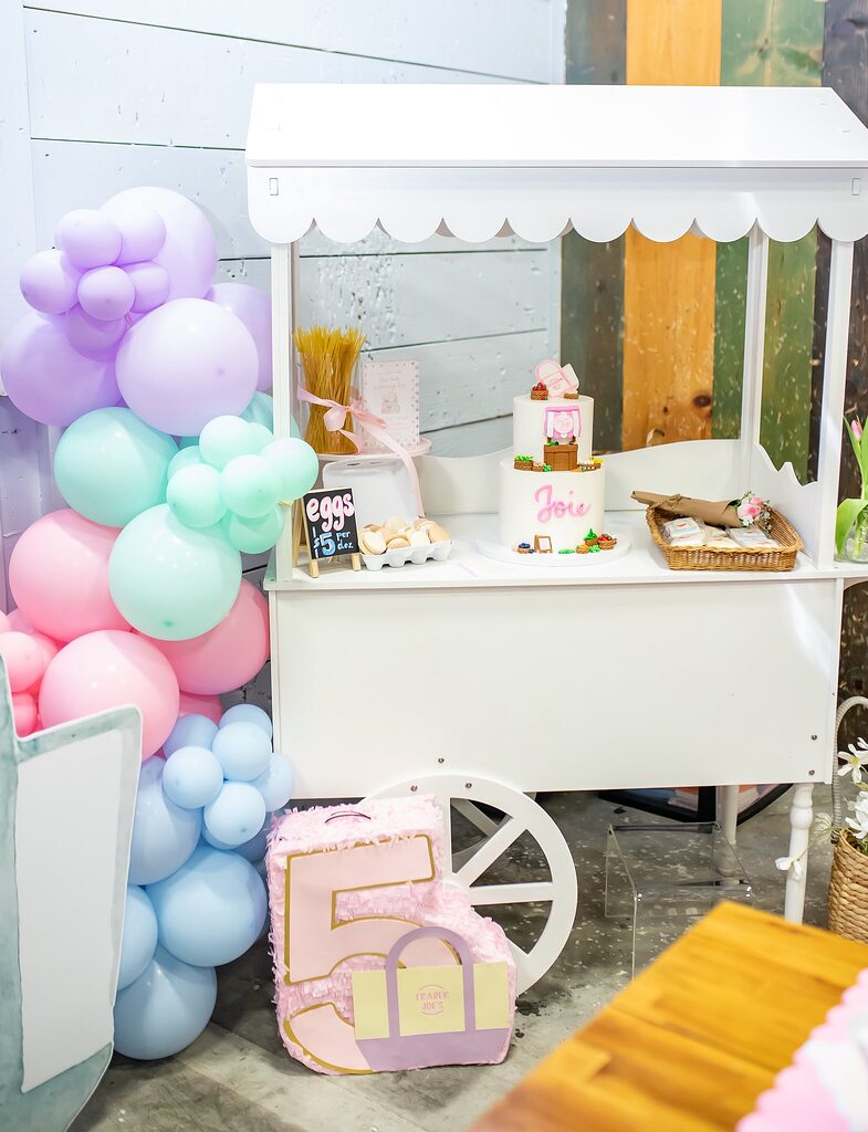 White party trolley display featuring a two-tier cake labeled Joie, pastel balloons, and a number five piñata.