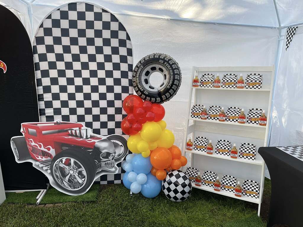 Hot Wheels themed party display with checkard backdrop, balloon arch, and party favors on a white shelf.