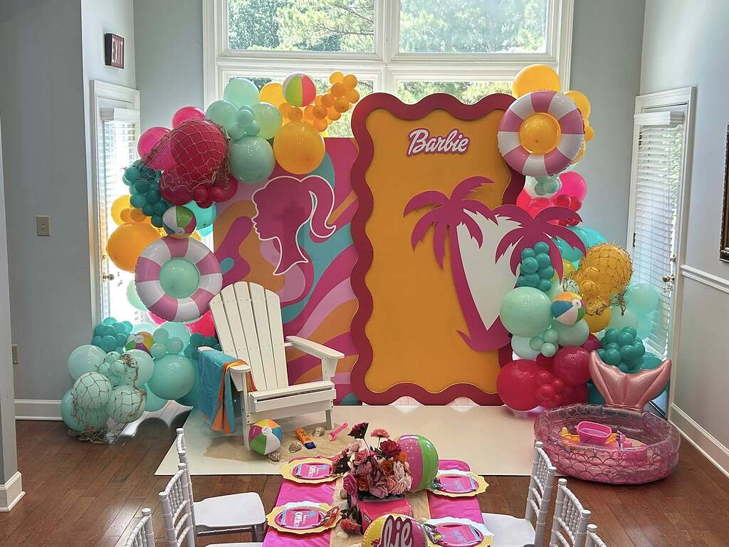 Barbie themed party photo backdrop featuring tropical decorations, a white adirondack chair, and vibrant balloons.
