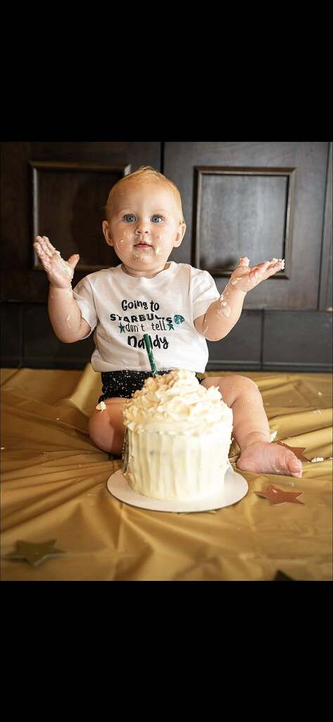 Baby covered in frosting sitting behind a white smash cake while wearing a Going to Starbucks shirt.