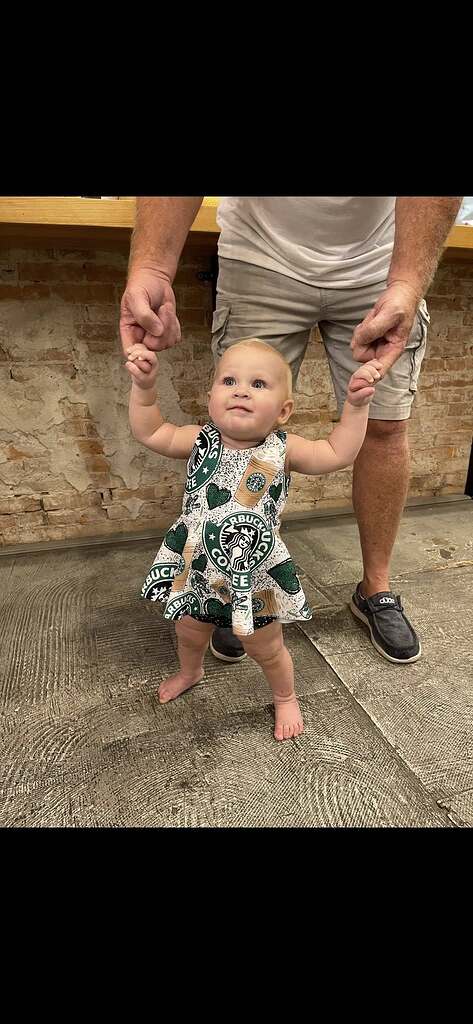 Baby girl wearing a coffee-themed print dress being helped to stand by an adult