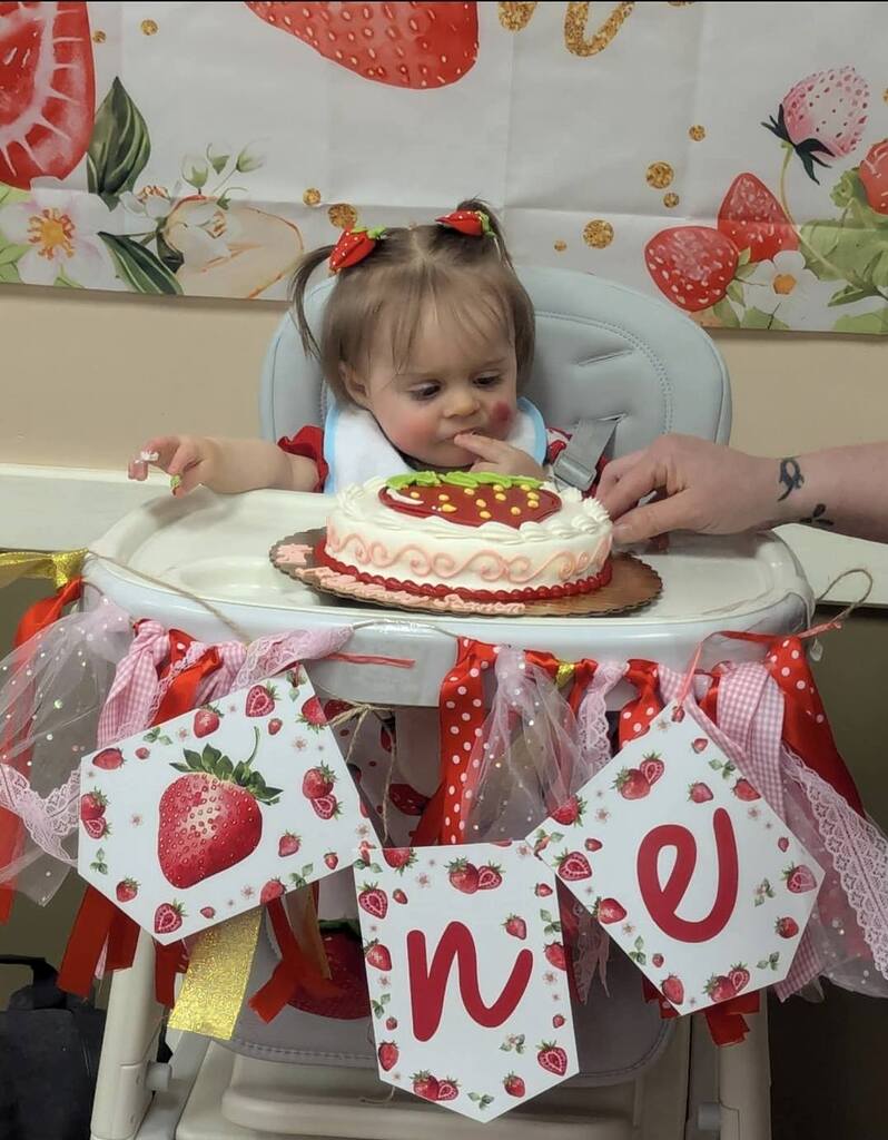 Baby celebrating first birthday in a high chair with a strawberry cake and strawberry-themed decorations.