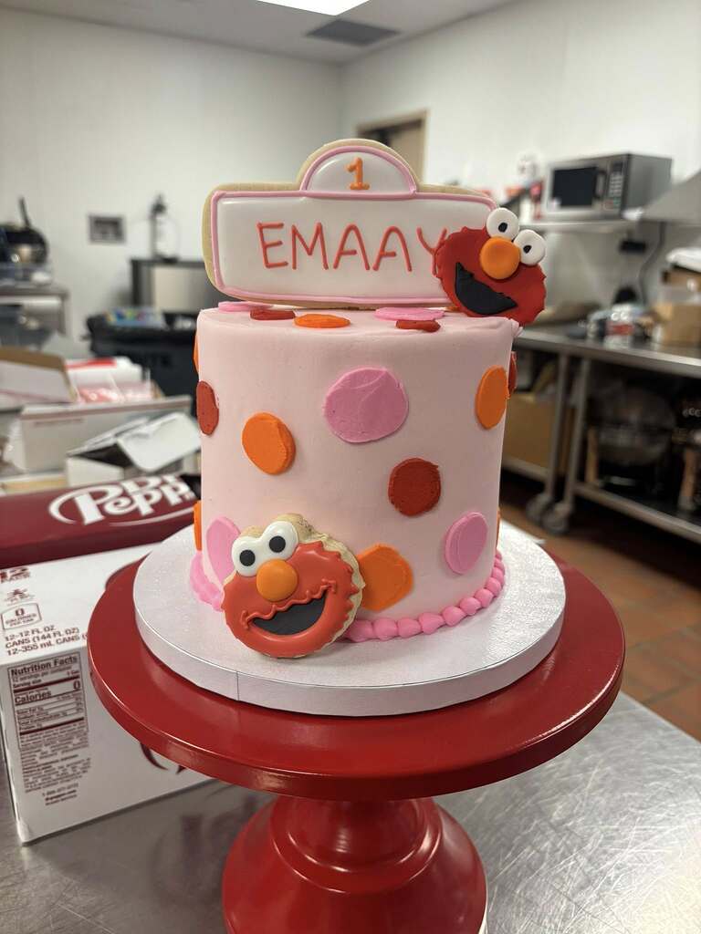 First birthday cake with pink frosting, colorful polka dots, Elmo decorations, and a name plaque reading Emaay.