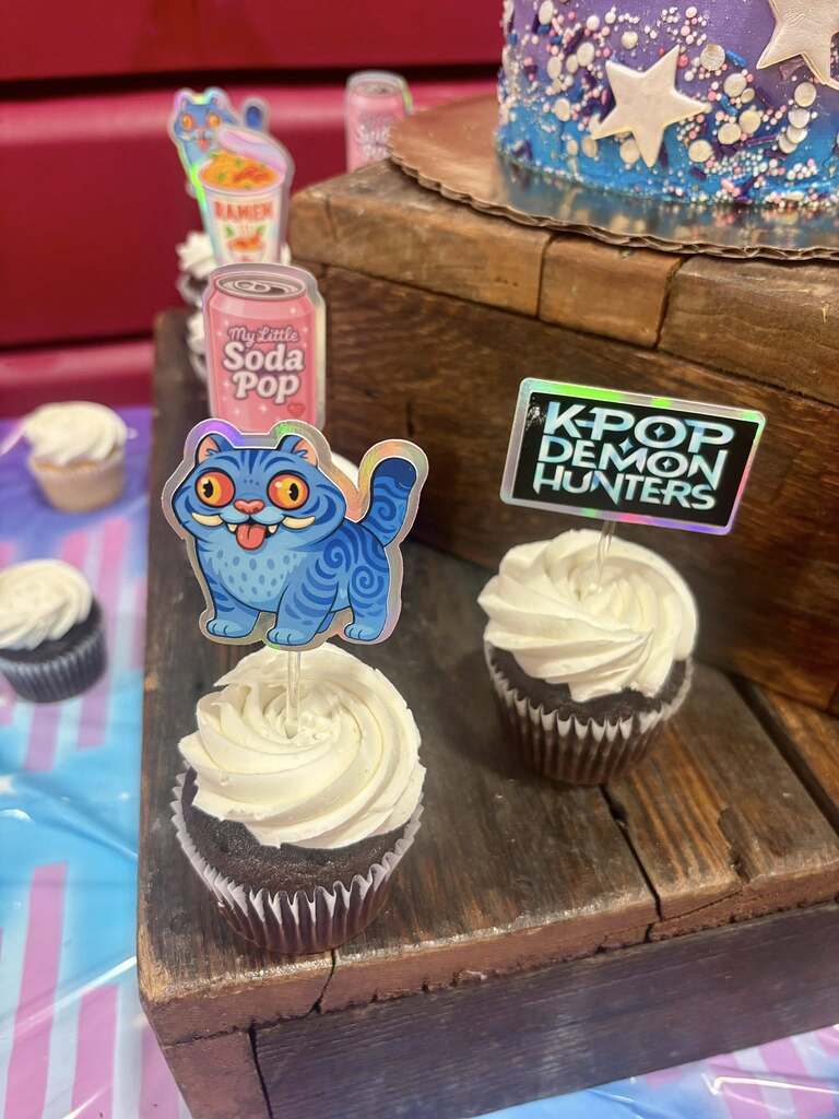 Cupcakes decorated with colorful holographic food and character toppers including a blue cat and K-pop sign.