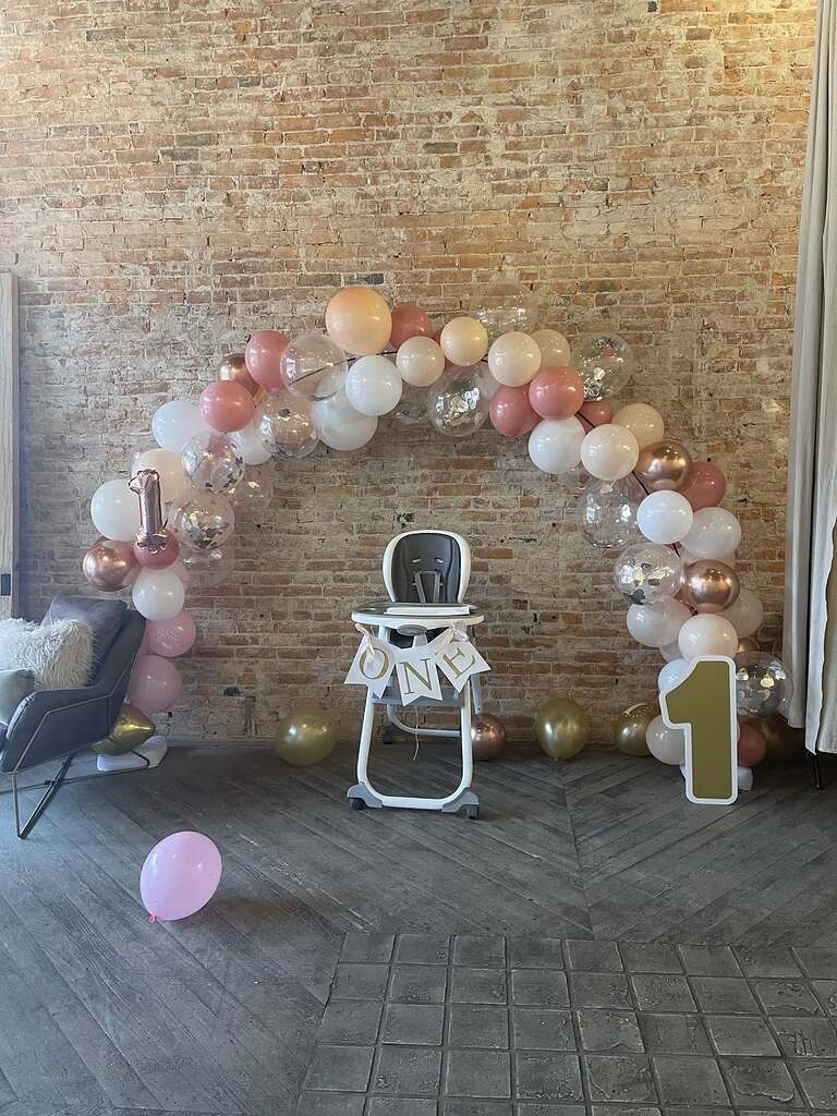 High chair for first birthday party decorated with a balloon arch and a One banner in front of a brick wall
