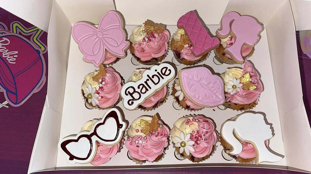 Box of twelve Barbie-themed cupcakes decorated with pink frosting, floral accents, and fondant cutouts.