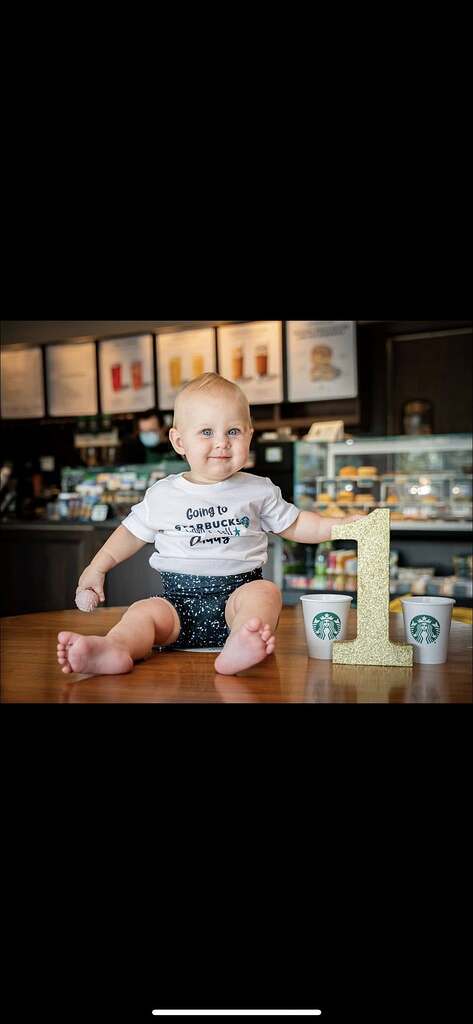 A one-year-old baby wearing a Starbucks-themed onesie, sitting at a table with a gold number one decoration.