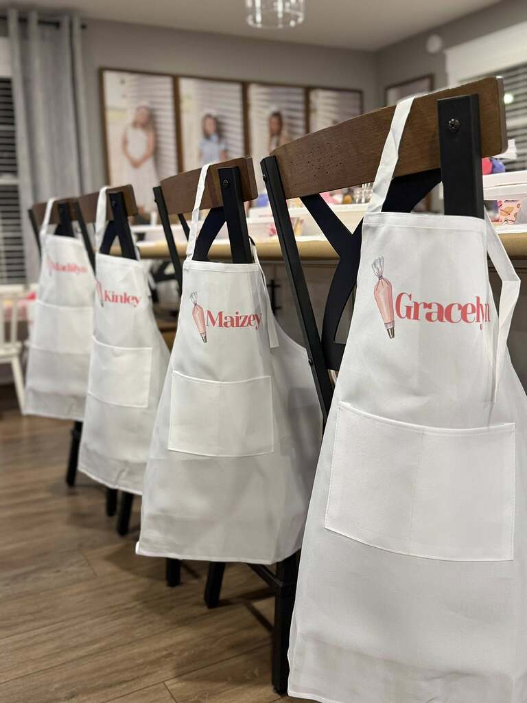Four white personalized baking aprons with piped icing design hanging on wooden chairs.