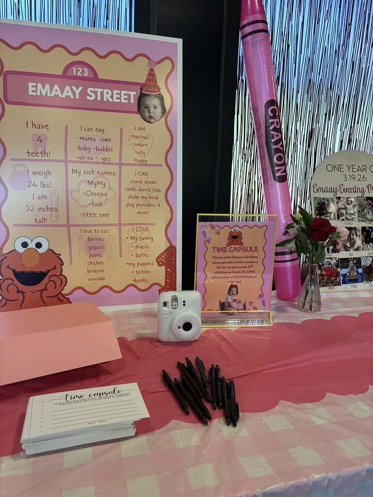 Table display for a first birthday party with milestone poster, time capsule sign, camera, and guest note cards.