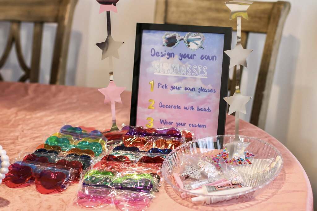 Table with a Design Your Own Sunglasses sign, plastic sunglasses in bags, and a bowl of beads for crafting.
