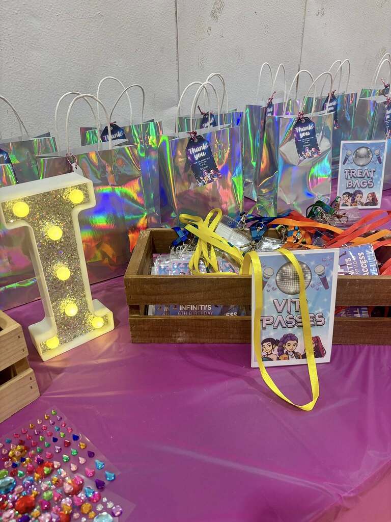 Table display for a themed birthday party featuring holographic treat bags, VIP passes with lanyards, and a light-up letter I