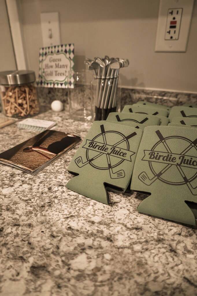 Stack of green koozies featuring a Birdie Juice golf logo with a guess the golf tees game sign in the background.