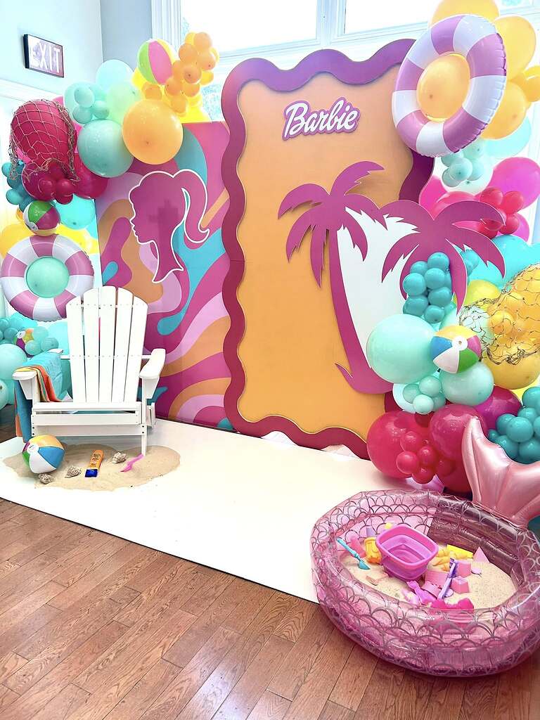 Barbie-themed beach party backdrop with balloons, palm tree, and white adirondack chair on sand