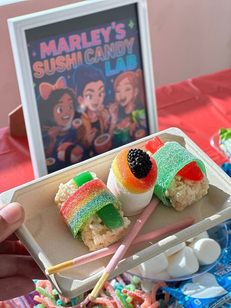 Hand holding a tray of handmade candy sushi treats featuring rice cereal rolls and colorful gummy toppings