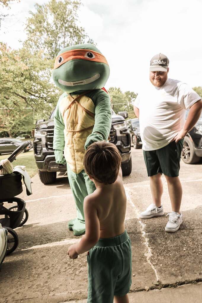A young child holding hands with a Teenage Mutant Ninja Turtle character mascot in a driveway.
