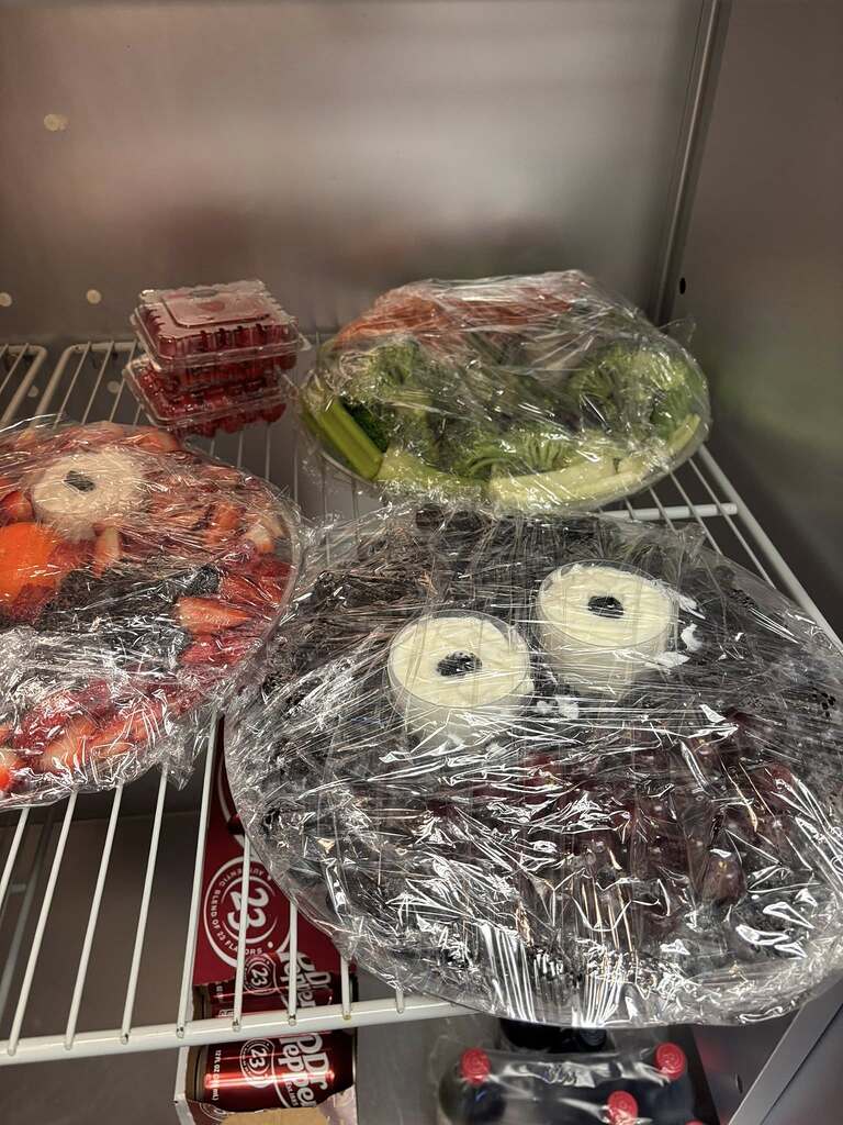 Plastic-wrapped fruit and vegetable platters with dip in a refrigerator shelf.