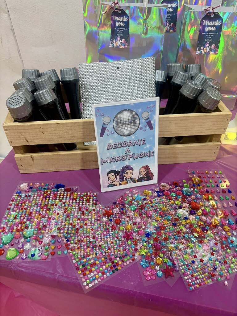 Table with a wooden crate of toy microphones and colorful adhesive rhinestone stickers at a DIY party station.
