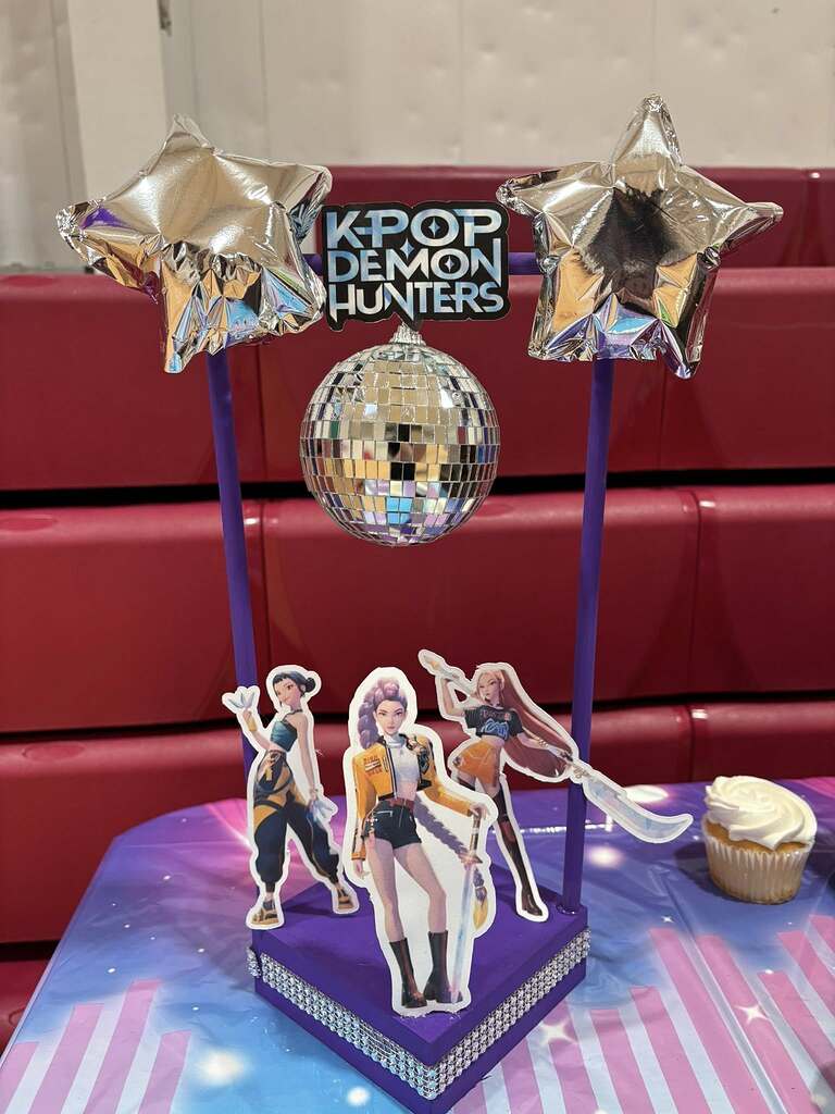 K-pop Demon Hunters party centerpiece with disco ball, star balloons, and cutouts of three characters.