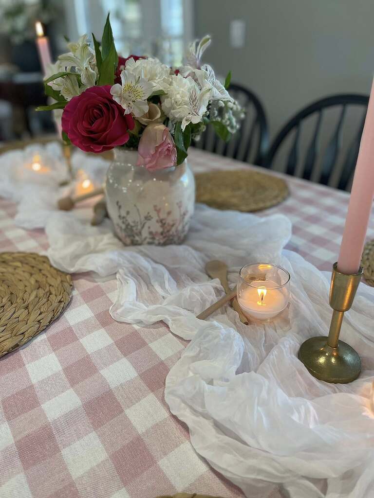 Dining table set for a special occasion with a pink checkered runner, floral vase, candles, and woven placemats.