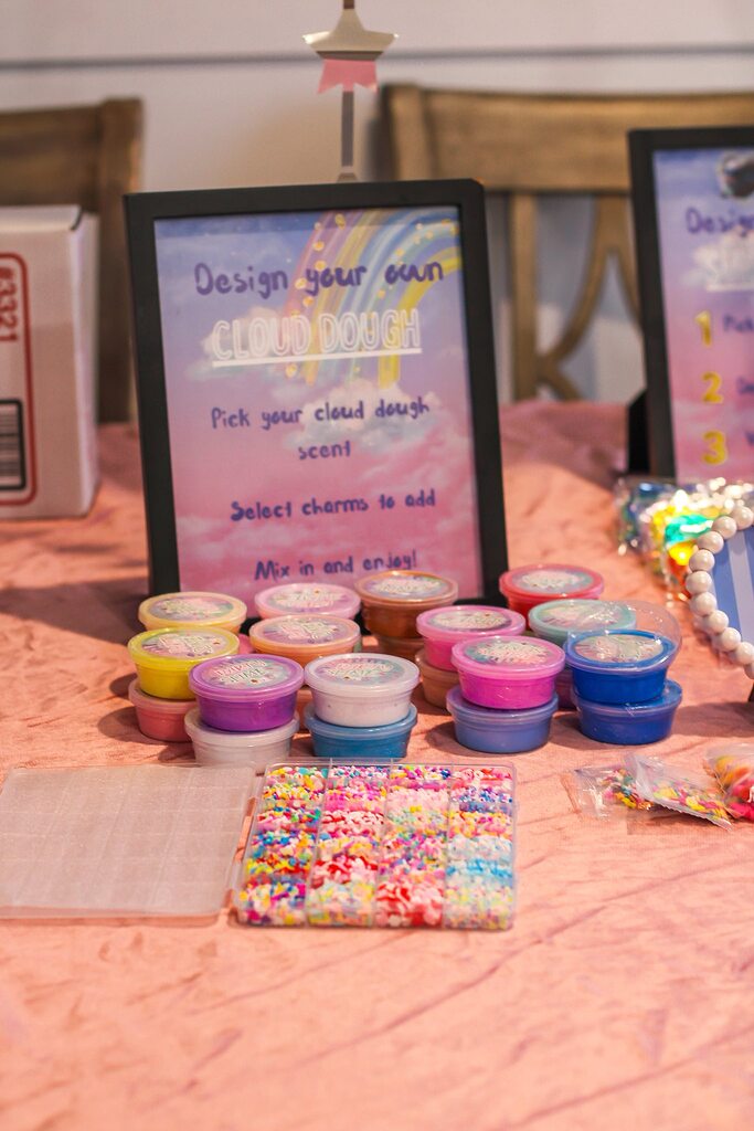 Cloud dough craft station with colorful dough pots, selection of mix-in charms, and a design instruction sign.