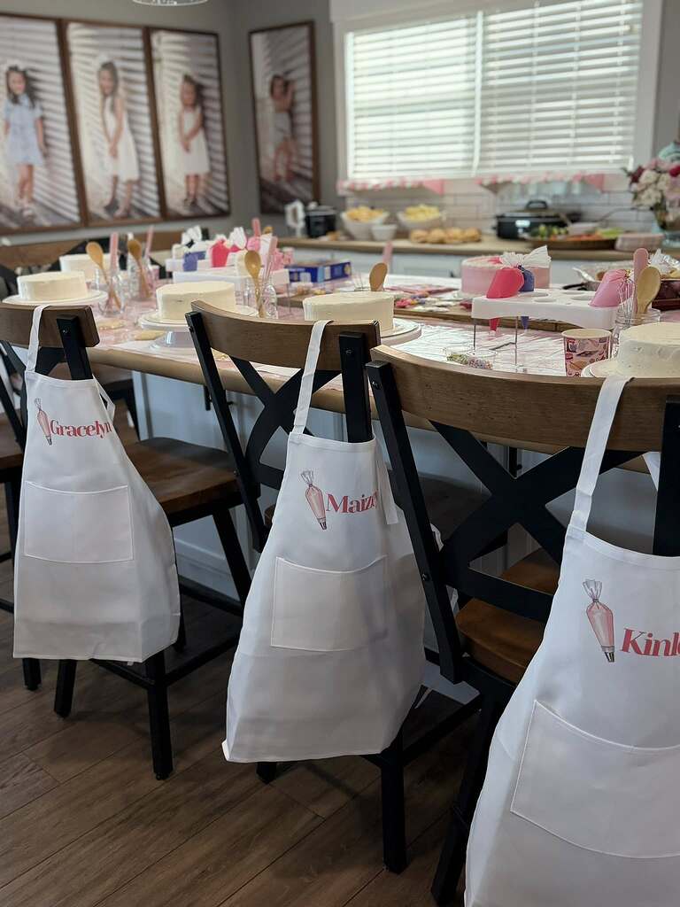 Personalized white children's aprons with names and piping bag graphics hanging on chair backs at a cake decorating party table