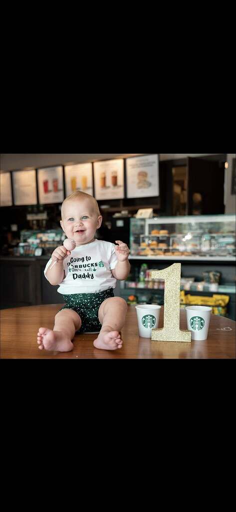 A one-year-old baby wearing a custom Going to Starbucks with Daddy white onesie sitting on a table with a number one prop.