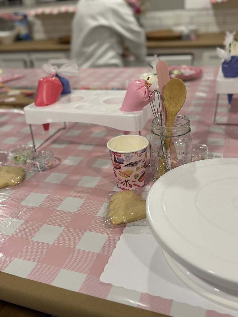 Decorated cookie party setup with piping bags, utensils in a mason jar, cookie cutters, and paper cups on a pink gingham tablecloth