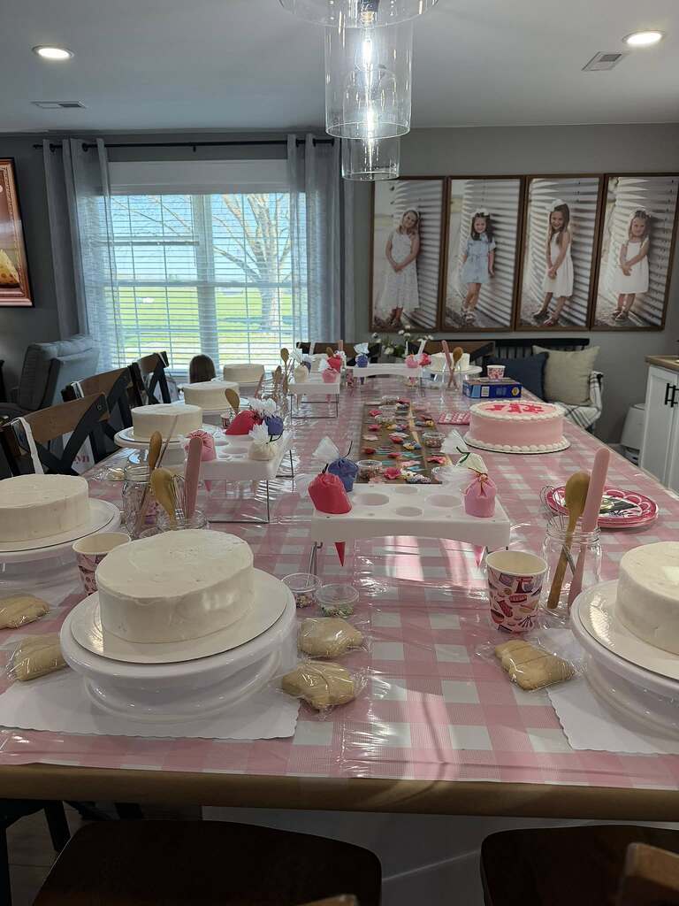 Dining table set up for a cake decorating party featuring multiple plain cakes, piping bags, and cookies.