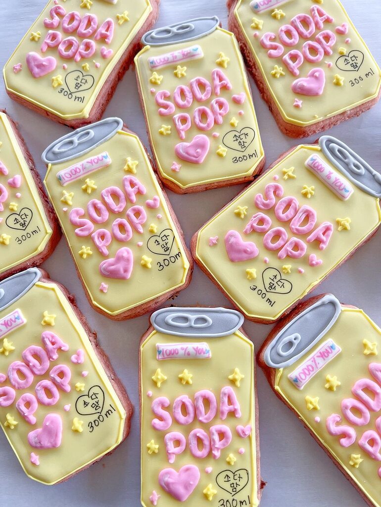 Group of soda can-shaped cookies decorated with yellow icing, pink text, and small hearts