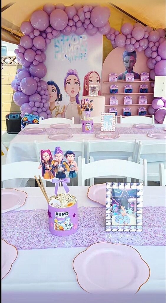 Table setting with Rumi themed party decorations, character centerpieces, lavender balloon arch, and display shelf.