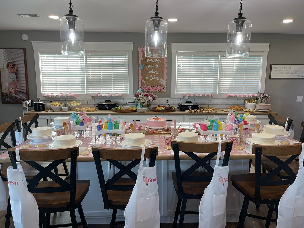 Table set for a cookie decorating party with personalized aprons, frosting bags, and blank frosted cakes.