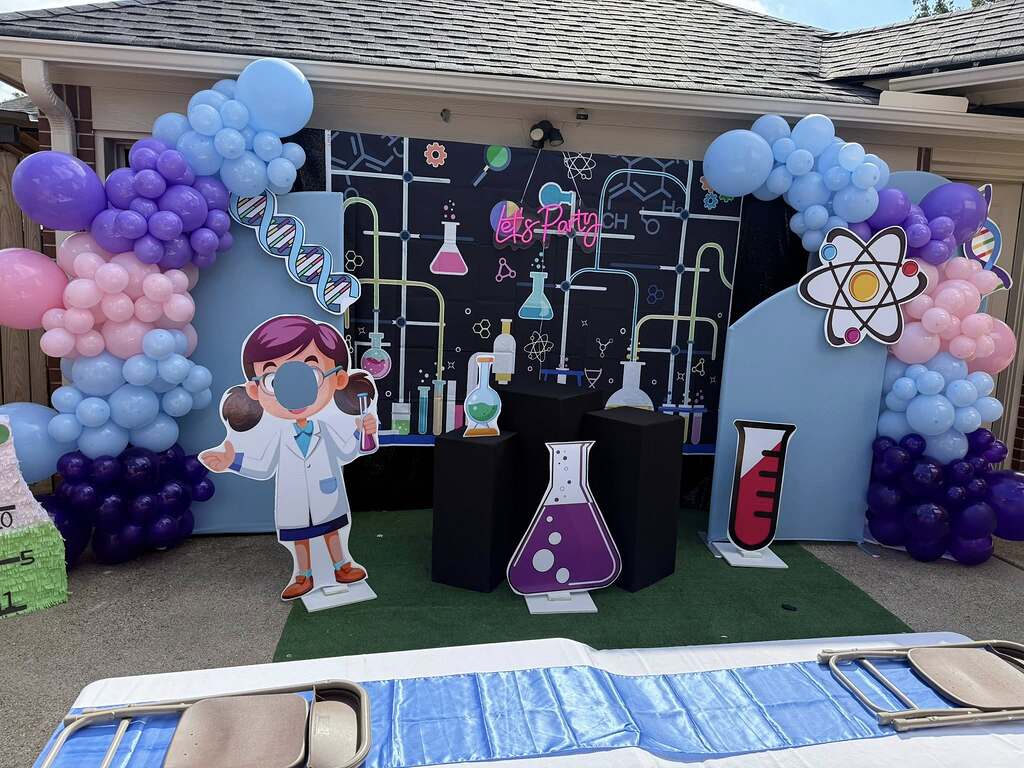 Mad scientist themed party backdrop with balloons, lab supply cutouts, and a character photo cutout.